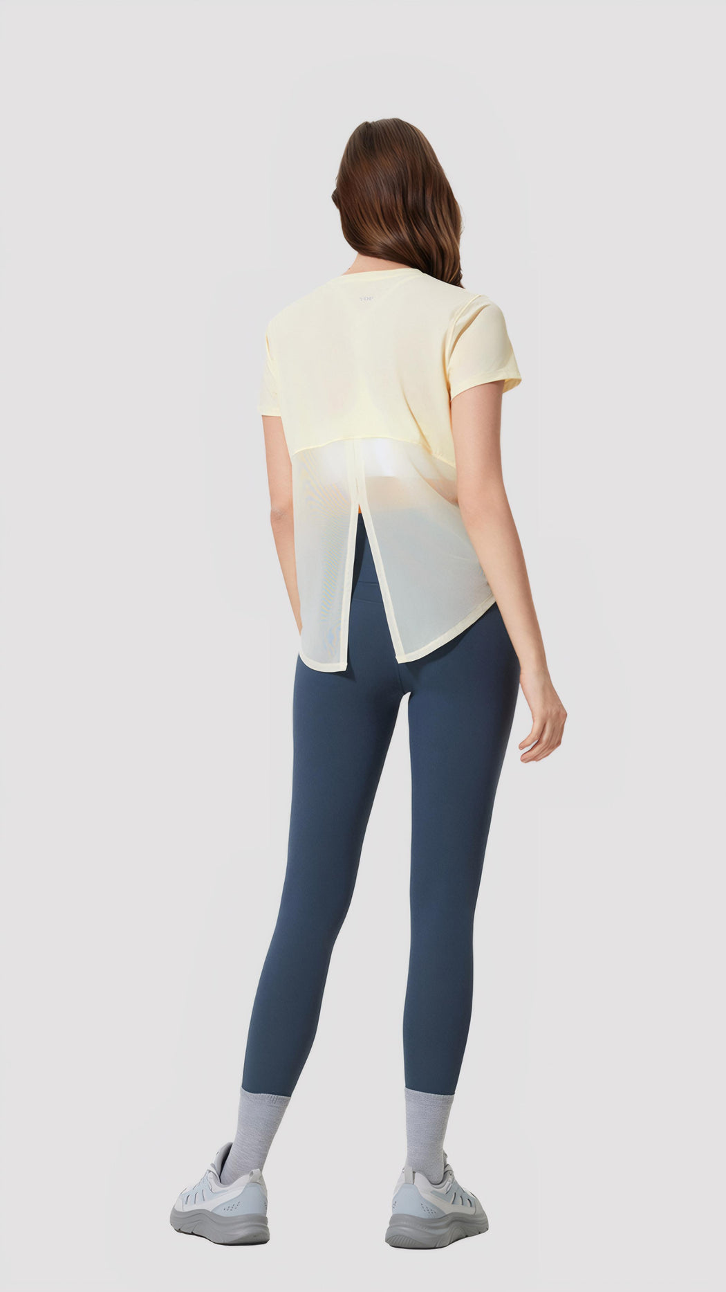 Woman wearing Aero Flow Active Tee in light yellow with back slit paired with blue leggings and sneakers, standing facing away